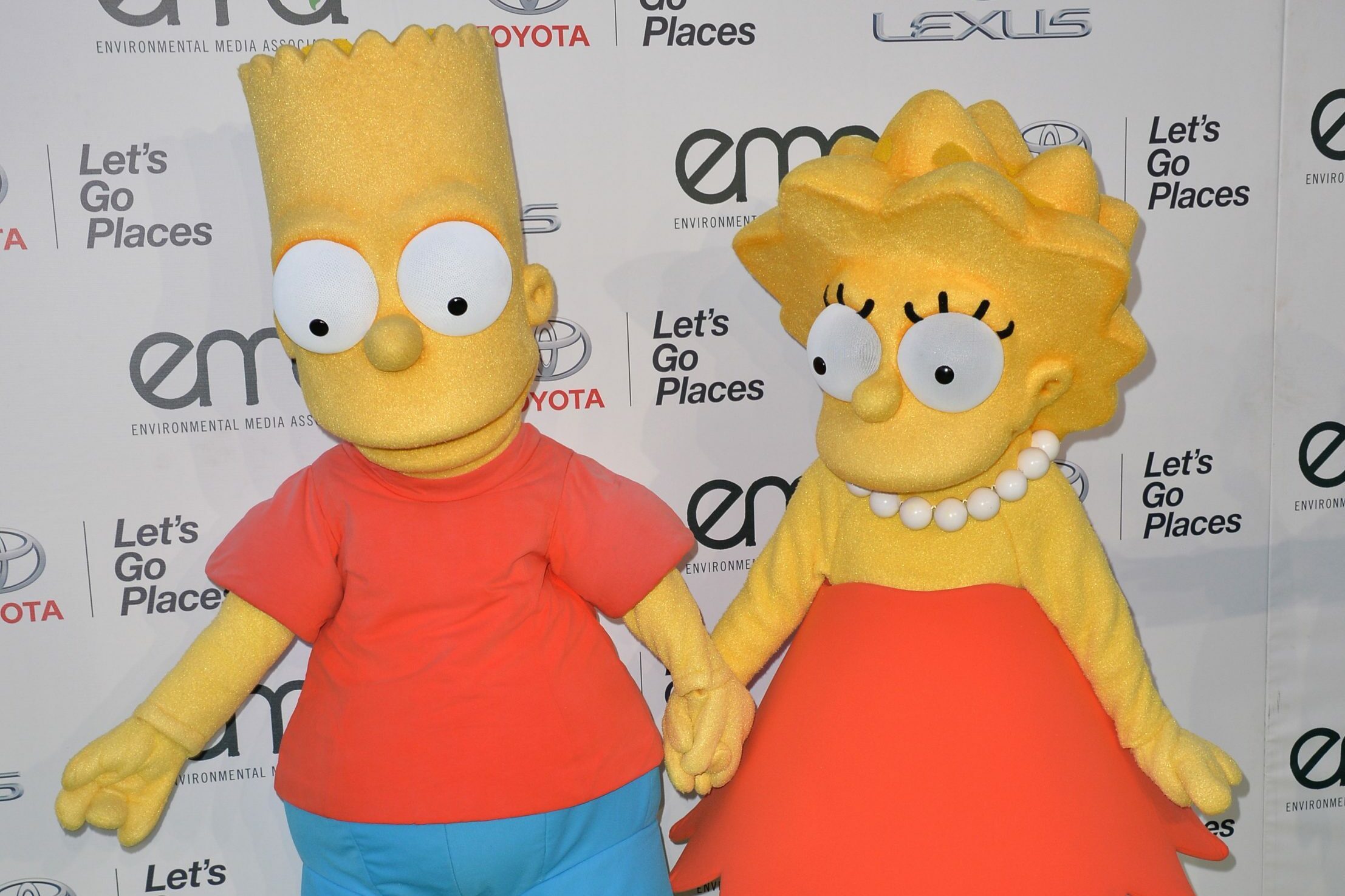 Two cartoon characters, holding hands, stand in front of a backdrop with "Let's Go Places" and "EMA" logos.