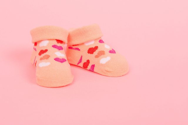 Peach-colored baby socks, decorated with colorful heart patterns, rest on a plain pink background.