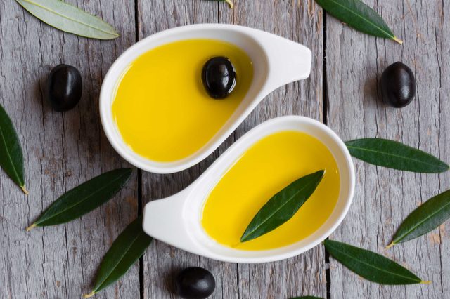 Two white dishes hold olive oil, one with an olive, the other with a leaf, set on weathered wood with scattered olives and leaves.