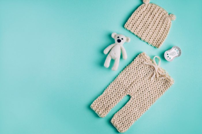 Baby accessories background: small hat and pants for newborn baby, dummy and bear toy over light green background with copy space; top view, flat lay