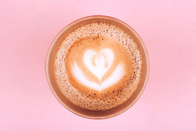 A latte cup displays heart-shaped foam art, set against a pink background.