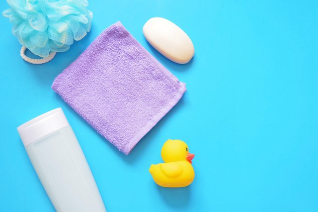 Bath essentials arranged on a blue background, including a blue loofah, purple towel, white soap, shampoo bottle, and yellow rubber duck.