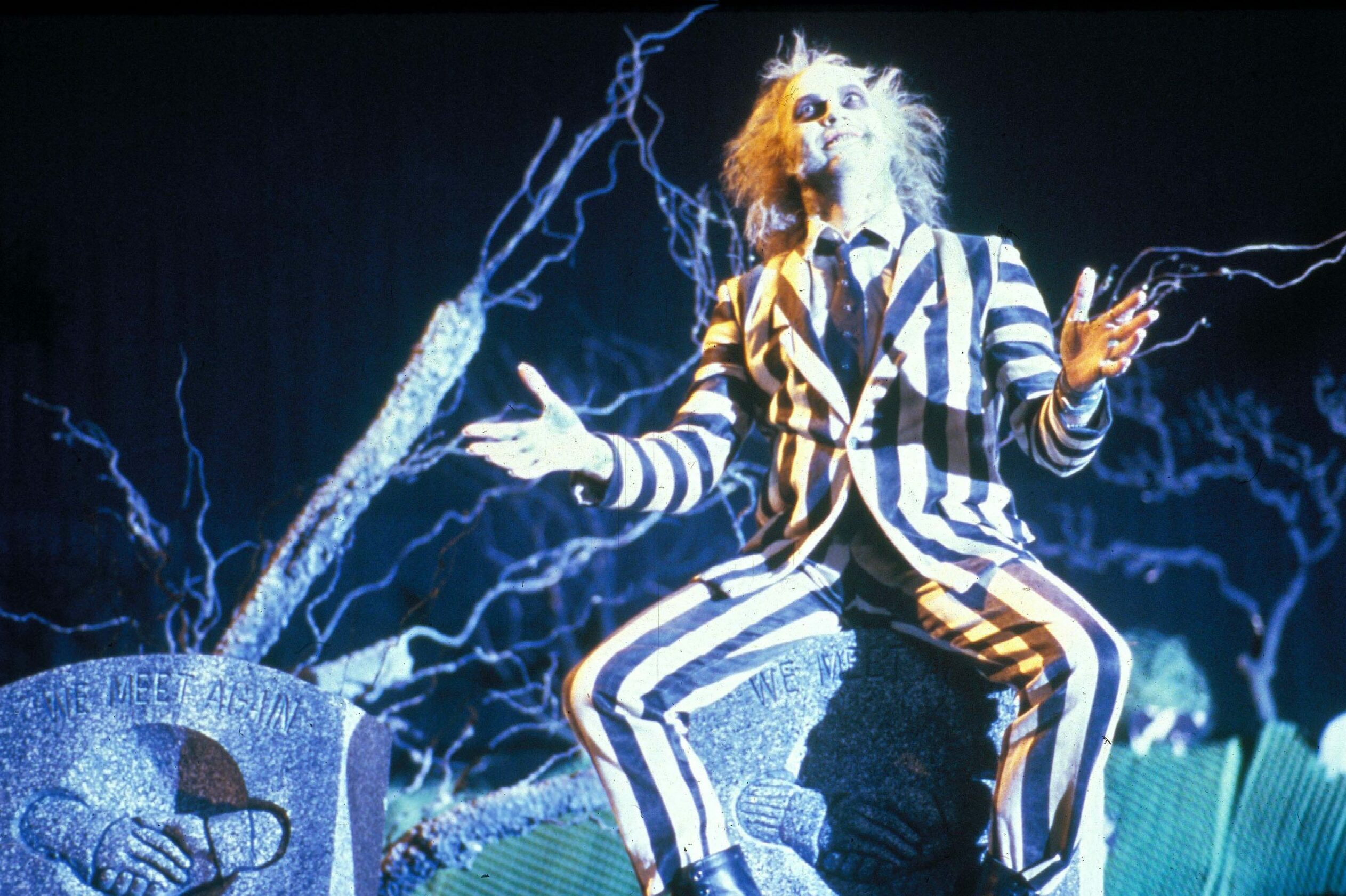 Individual dressed in stripe suit gesturing with open arms sits on tombstone in eerie, dark environment with twisted tree branches and gravestones.