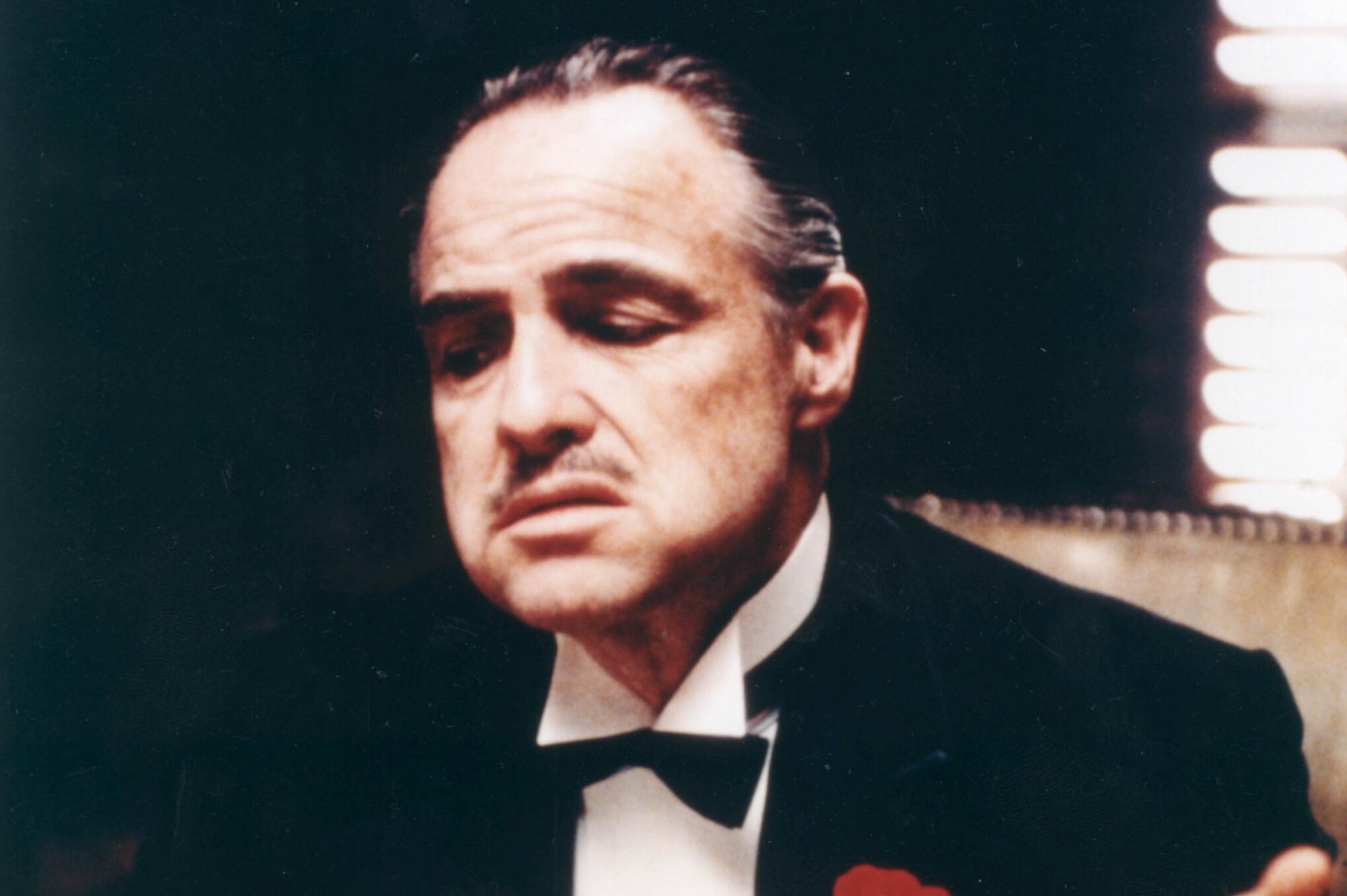 Man sits solemnly in a dark room, wearing a tuxedo with a red rose on the lapel, light filtering through blinds.