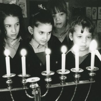 Children observe candles on a menorah, one child holding a candle, in a classroom decorated with artwork.