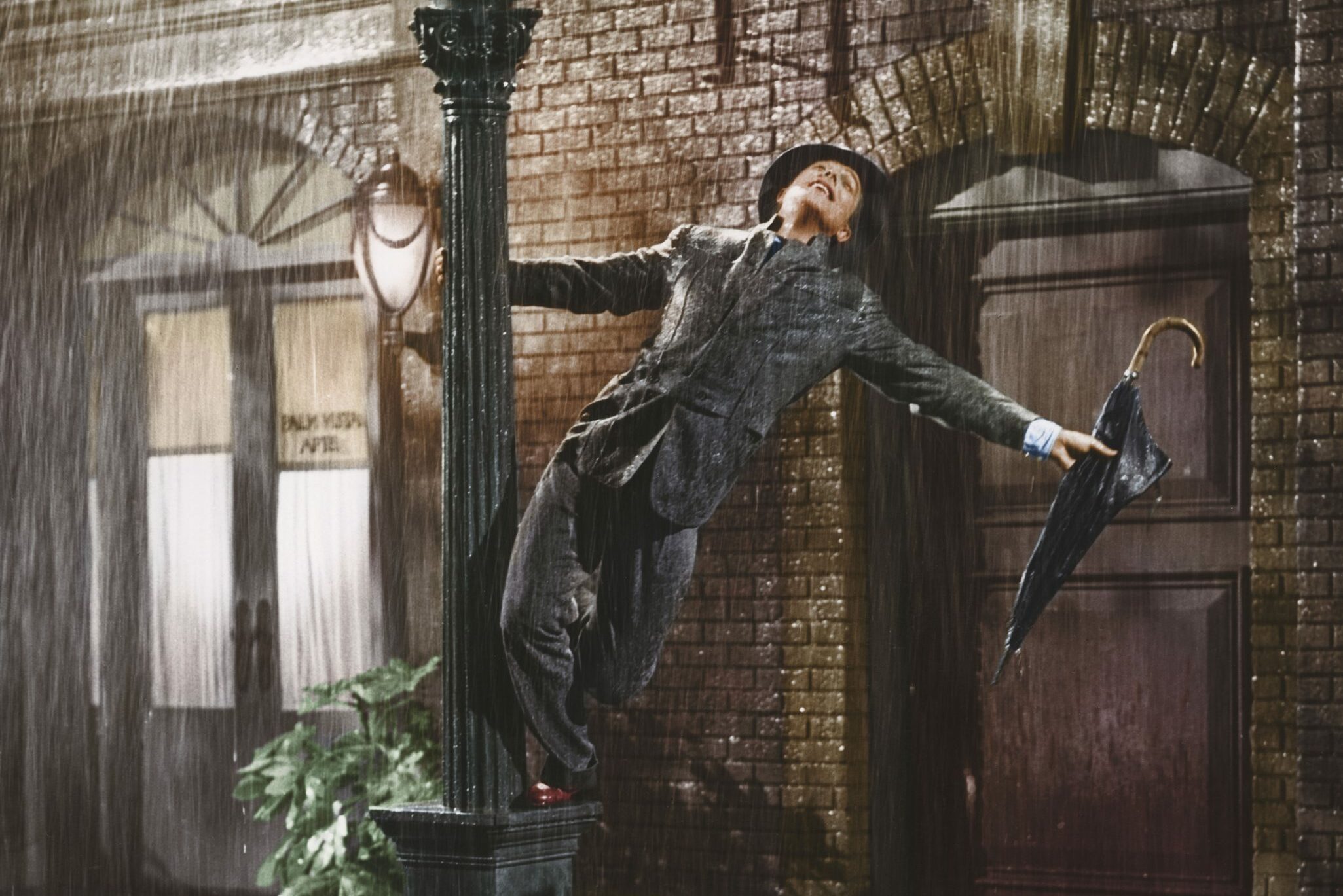 A person joyfully swings from a lamppost in the rain, holding a closed umbrella, against a brick building backdrop with large arched windows.