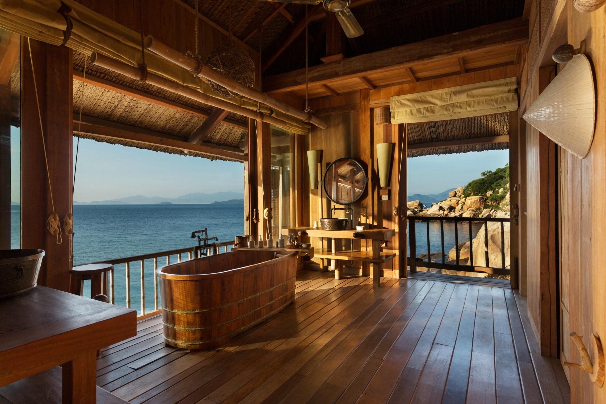 Wooden bathtub sits beside a sink in an open-air, rustic bathroom overlooking the ocean with mountains and rocks in the distance.