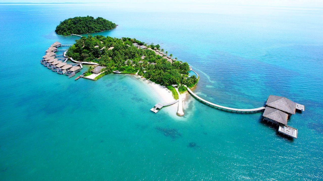 Aerial view of tropical island resort, surrounded by turquoise water, with villas, palm trees, sandy beaches, and a long curved wooden pier.