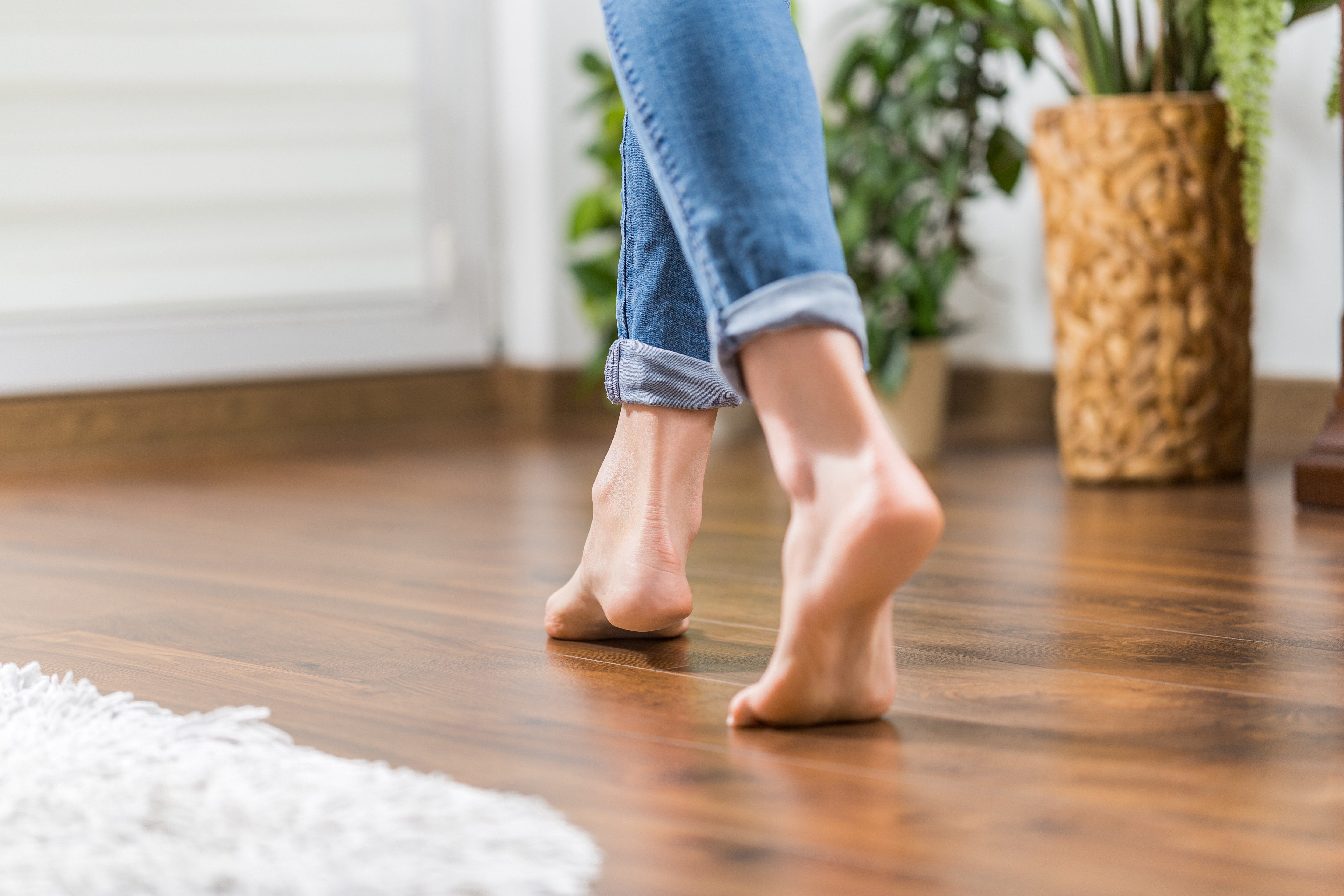 Bare feet tiptoe on wooden floor; person wears rolled-up jeans. Indoor plants and a textured rug enhance the cozy room setting.