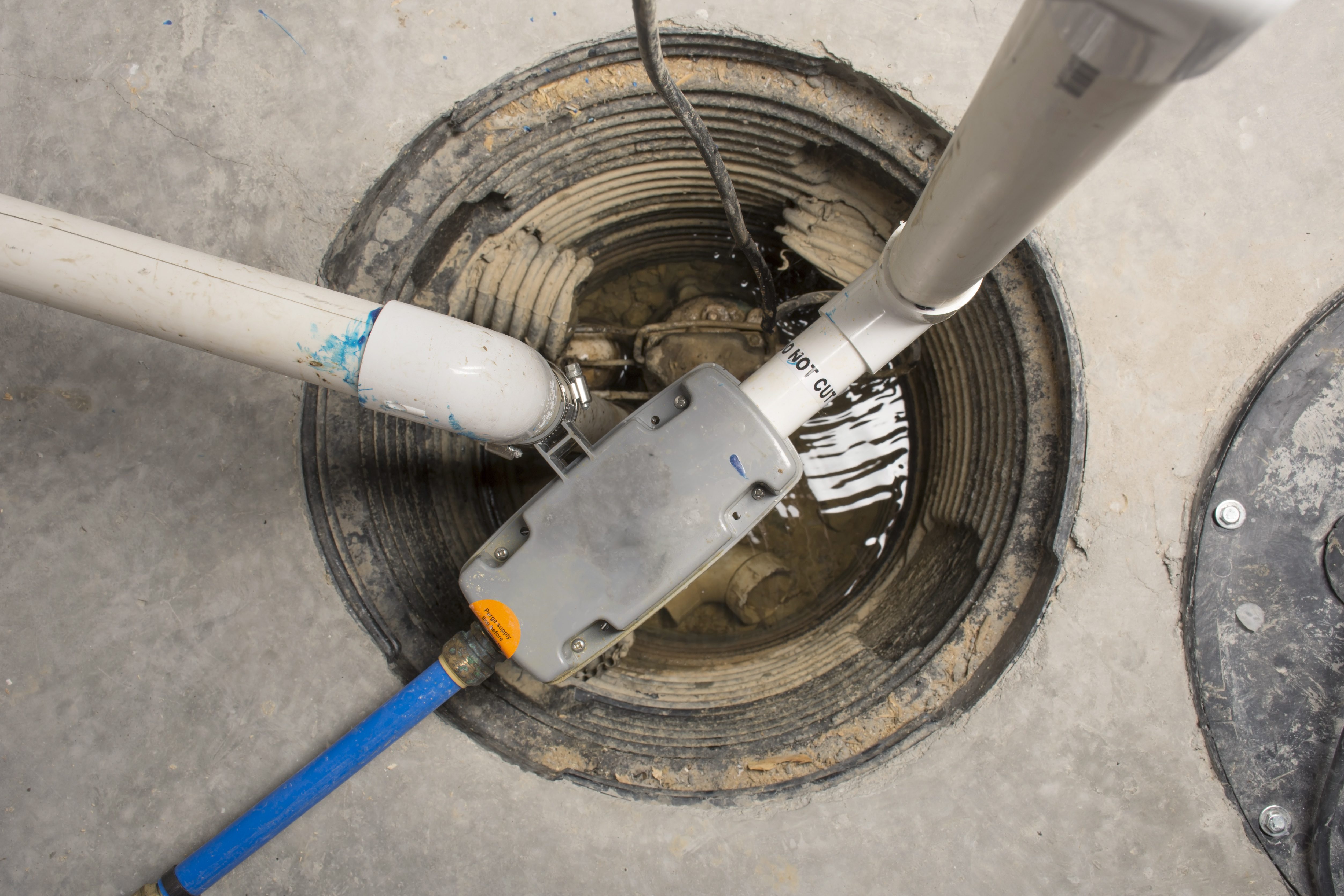 Pipes connect within a sump pit containing water and dirt, surrounded by a concrete floor. A label reads "DO NOT CUT."