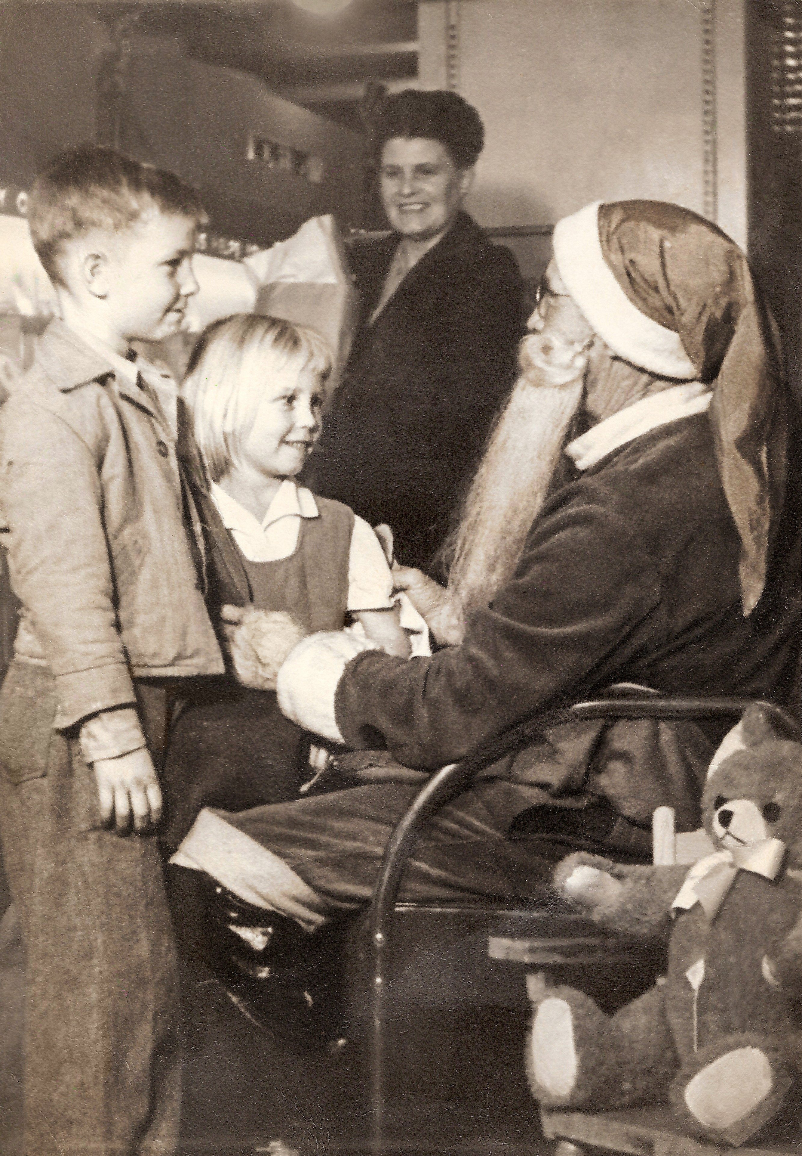 Santa Claus sits, talking to two children, in a cozy, festive setting with a smiling woman and teddy bear nearby.