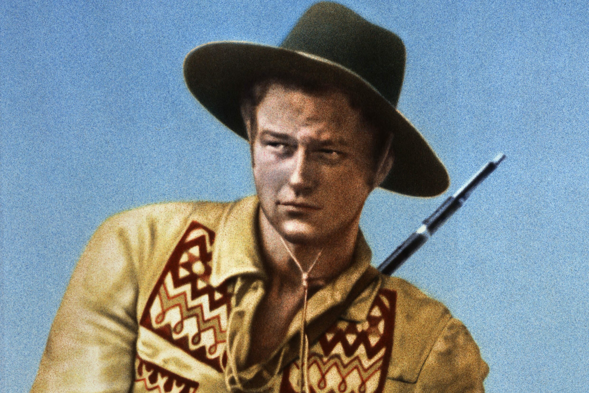 A man wearing a decorative yellow shirt and green hat holds a rifle. He poses against a clear blue sky, looking intently to the side.