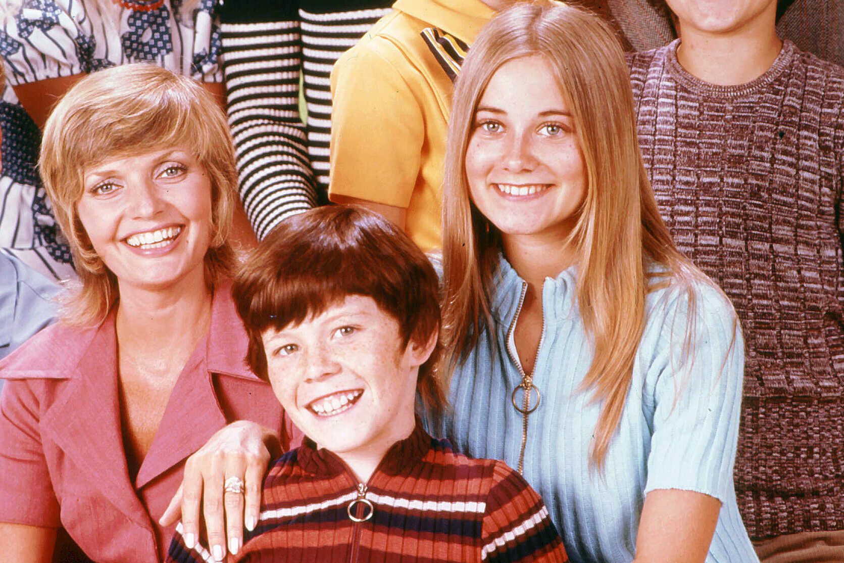 Three smiling people, sitting closely in colorful 70s-style clothing. The background features more individuals, partially visible, in patterned and striped attire.