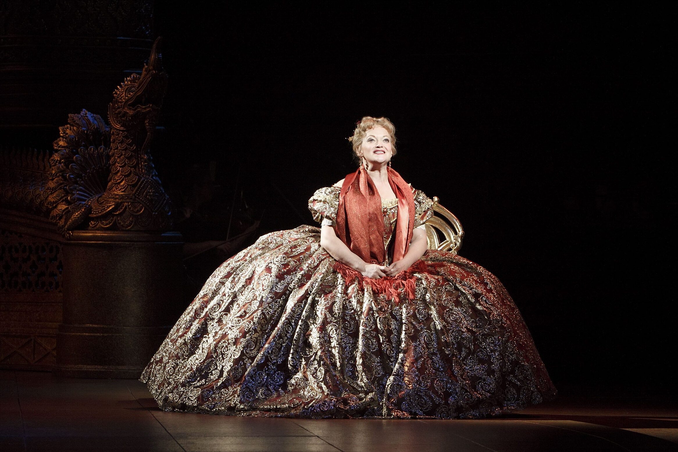 Woman sits elegantly, wearing a lavish, ornate gown with a red scarf in a dimly lit, theatrical setting, evoking a regal atmosphere.