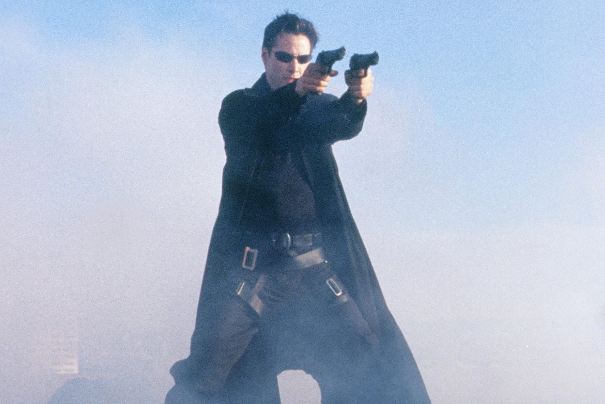 A person in a long coat aims two pistols forward, standing amidst foggy surroundings and wearing sunglasses.