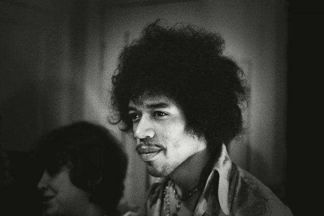 A person with curly hair looks forward; wearing a patterned shirt and necklace in a dimly lit room.
