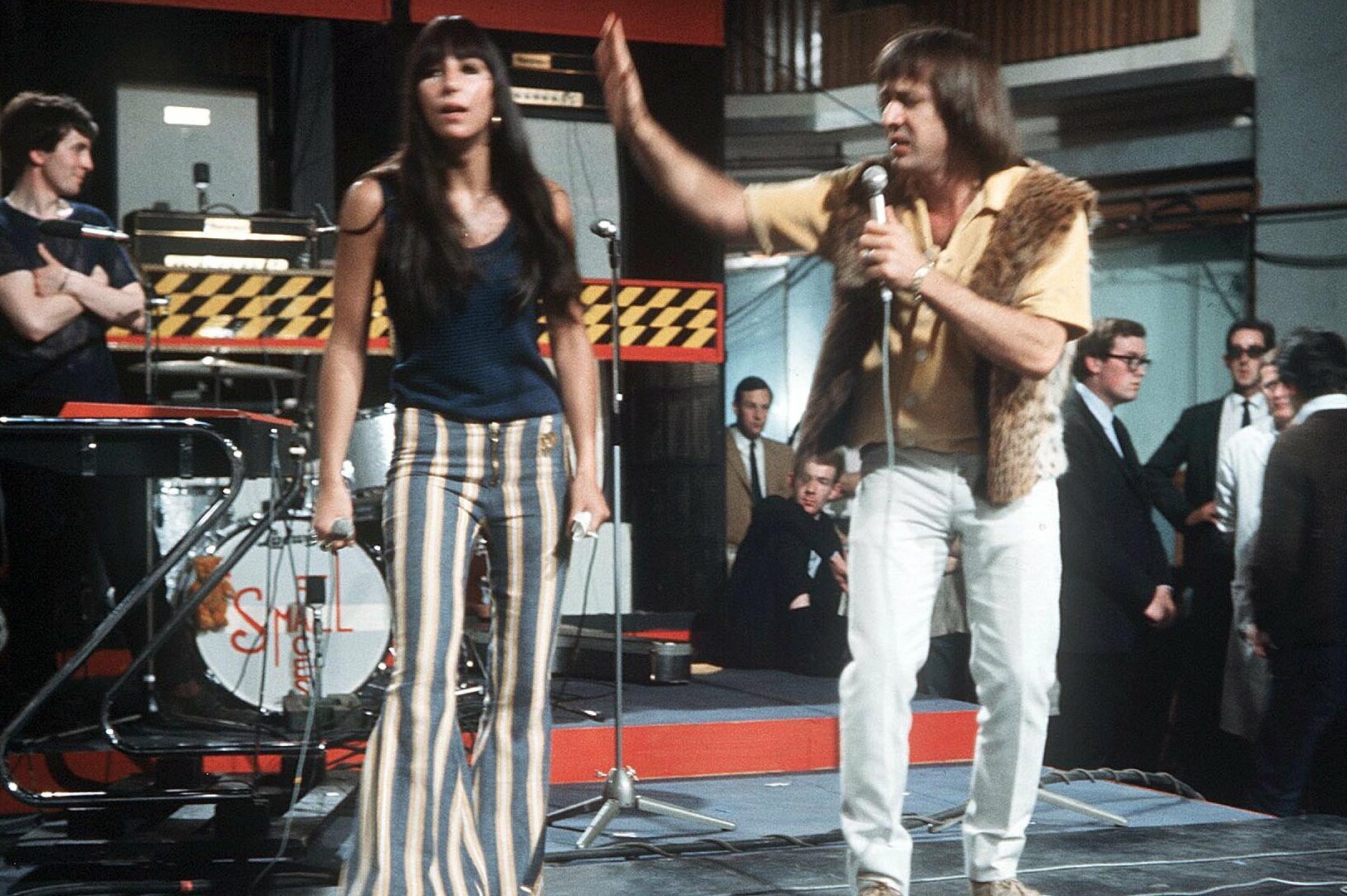 Two people are singing on stage. One holds a microphone, the other gestures animatedly. A band and audience are in the background.