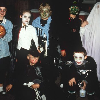 Children in Halloween costumes stand and crouch, posing together with face paint and masks in a dark outdoor setting. One holds a pumpkin.