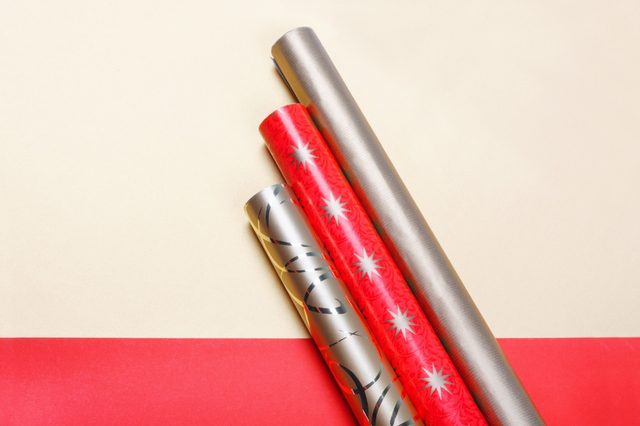 Three rolls of wrapping paper lie on a cream surface; designs include red with stars, silver with swirls, and plain gray.