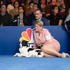 Dog lies beside woman, both resting on blue carpet, surrounded by spectators and NBC logo background.