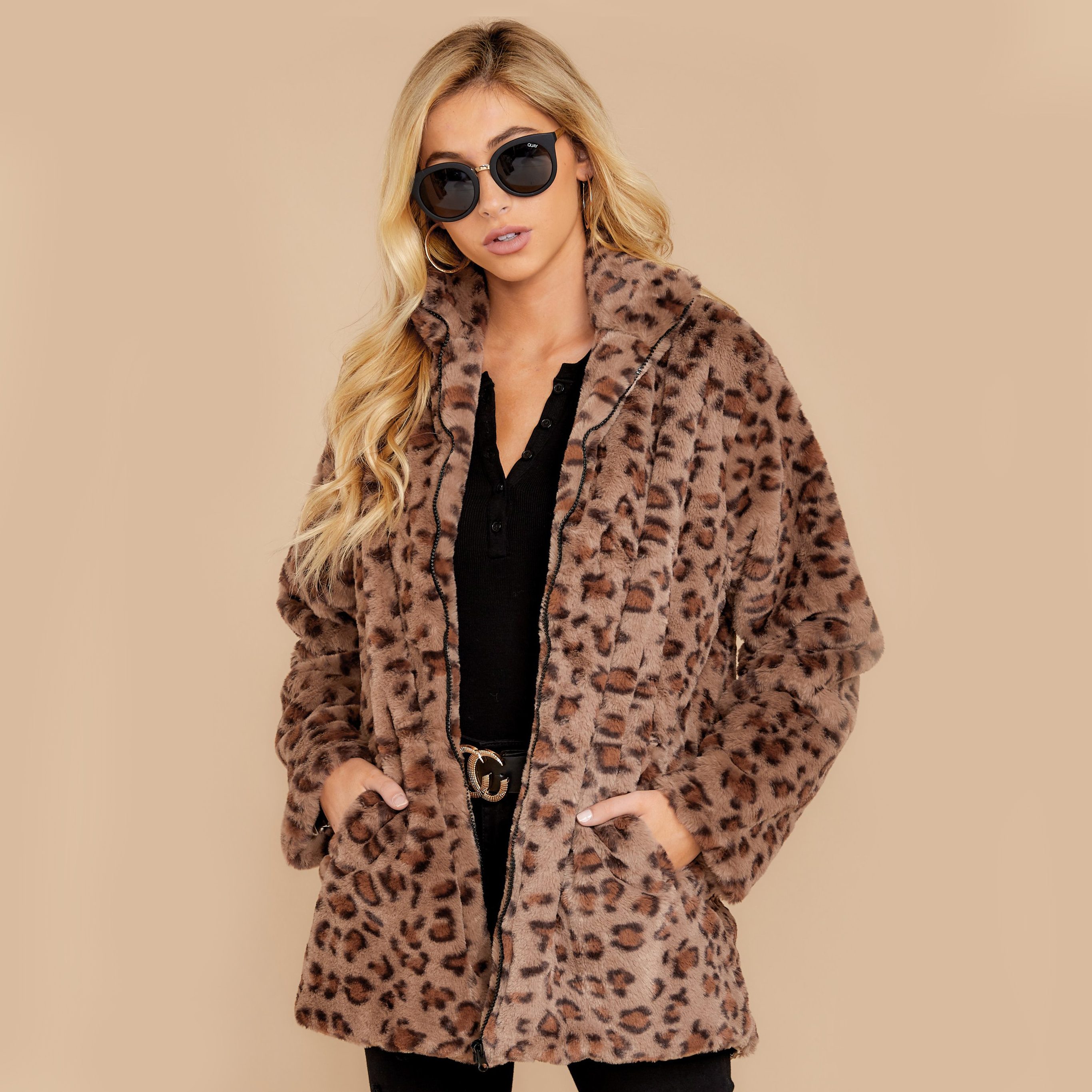 Woman wearing leopard-print coat and sunglasses, standing confidently against a plain beige background.