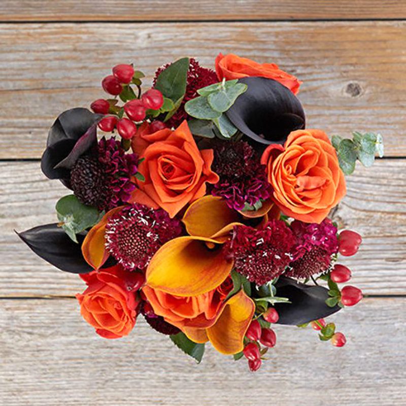 Bouquet featuring orange roses and dark calla lilies, arranged with red berries and leafy greens, placed on a rustic wooden surface.