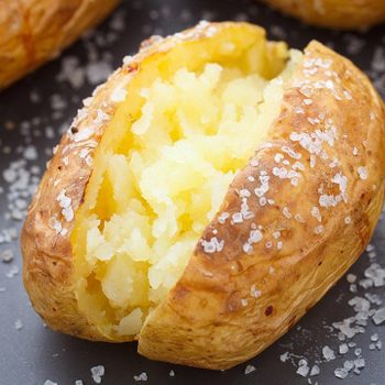 Baked potato rests open, displaying fluffy interior, surrounded by sprinkled coarse salt on a dark surface.