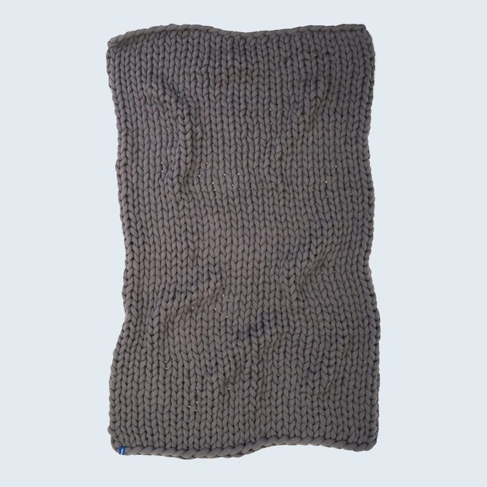 Chunky knit blanket lays unfolded, showcasing thick, braided patterns. The background is a plain light color.