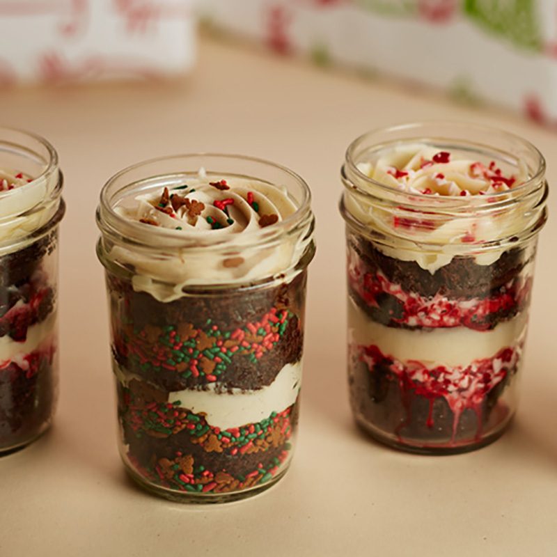Jar desserts alternate cake and frosting layers, adorned with festive sprinkles. They sit on a neutral surface with blurred holiday-themed background elements.
