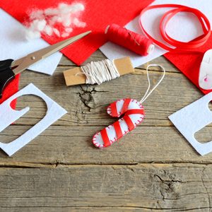 Scissors lie on felt sheets, crafting materials with candy cane ornament, thread, and stuffing on a wooden surface.