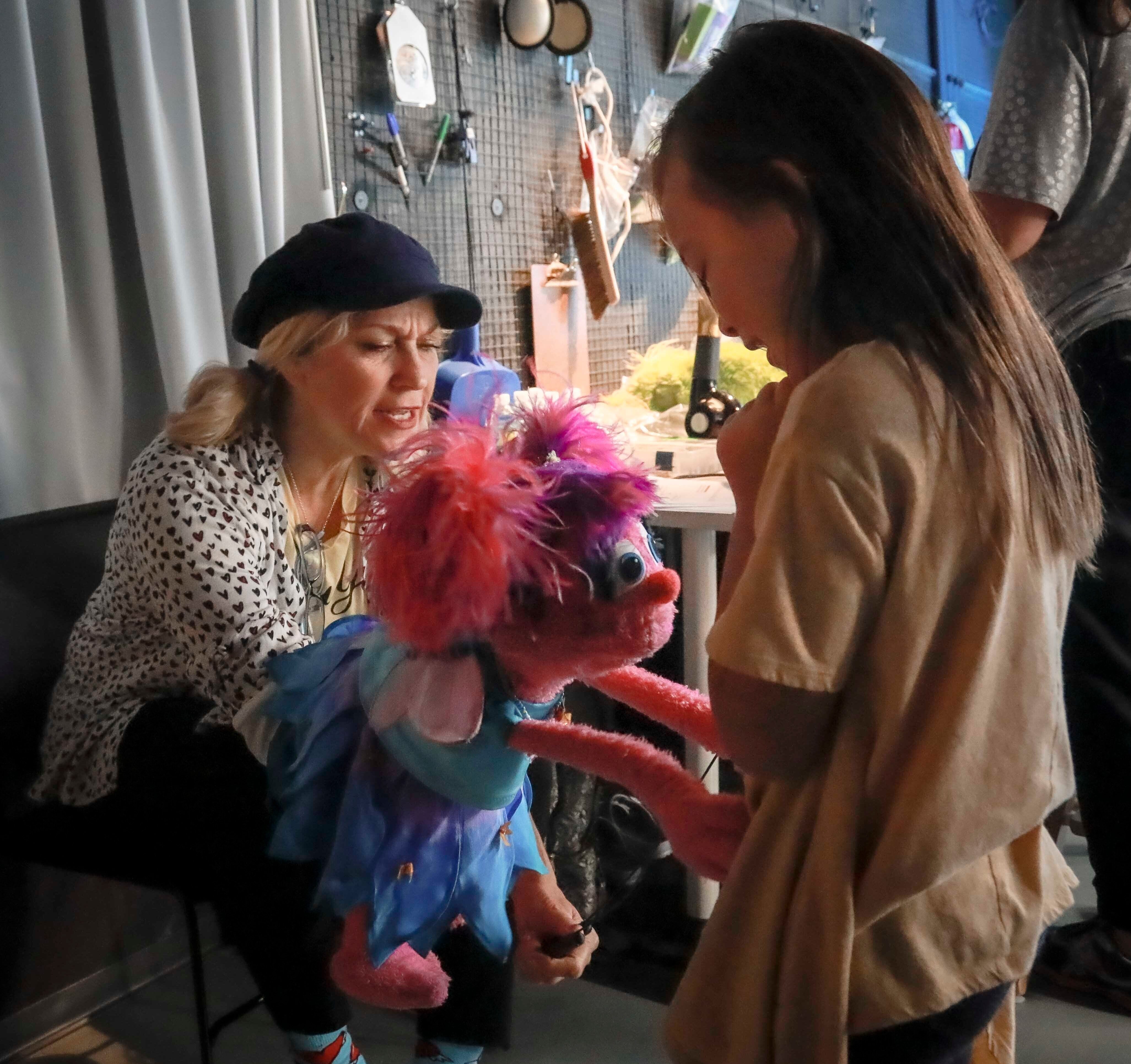 A puppet interacts with a child backstage, amidst a cluttered workbench and hanging tools in the background.