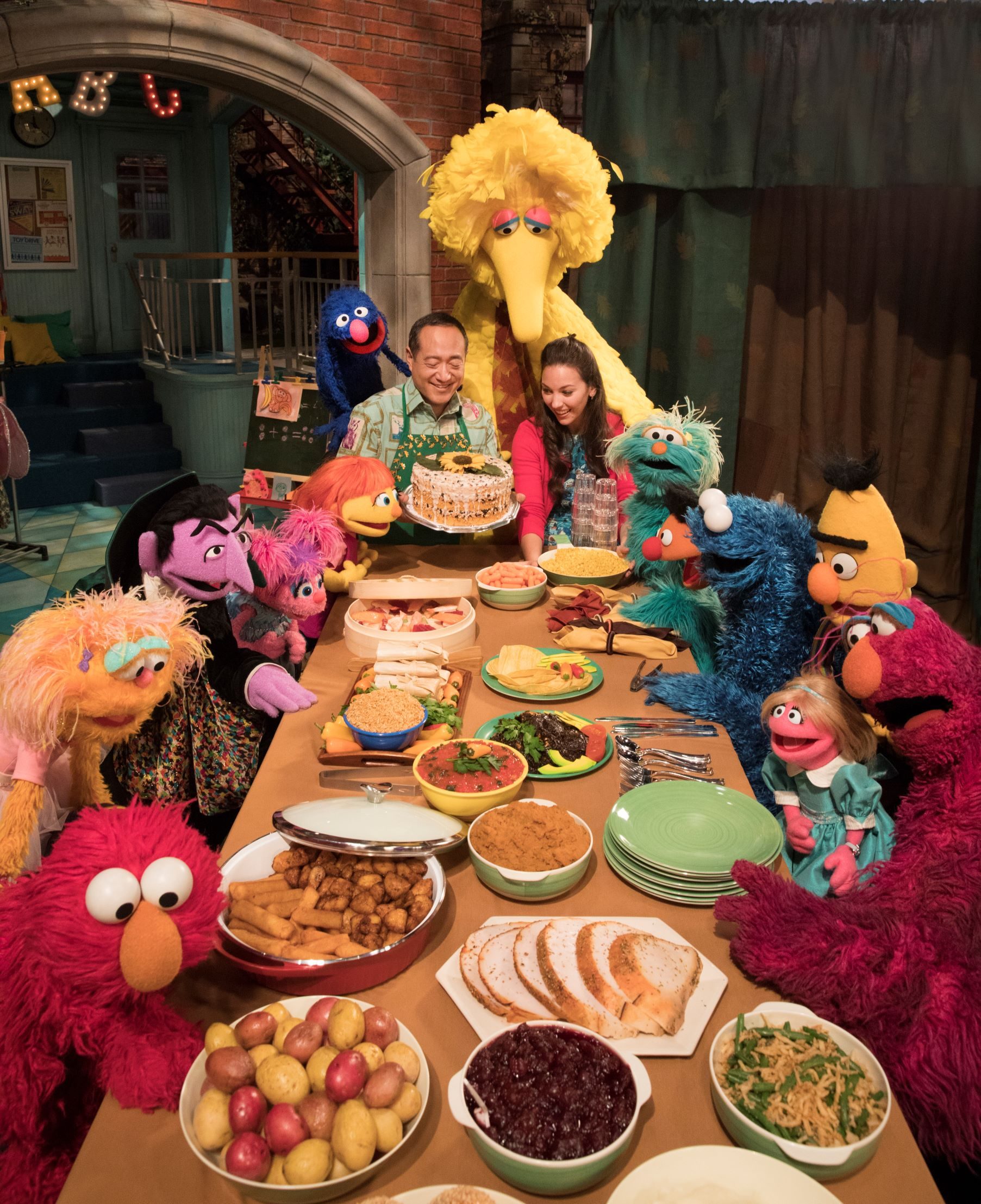 Puppets and two people gather around a table filled with diverse foods, celebrating in a warmly lit, inviting room.