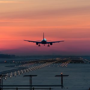 Airplane descends towards runway, glowing lights lining the path, with a vibrant sunset sky creating a colorful backdrop.