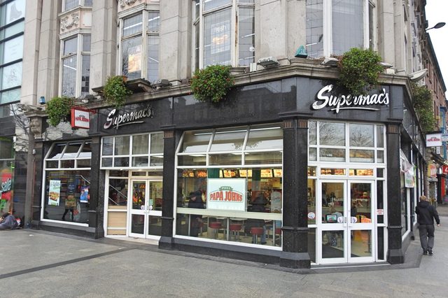 Restaurant storefront displaying "Supermac's" and "Papa John's" logos, situated on a busy city street corner with large windows and decorative stonework.