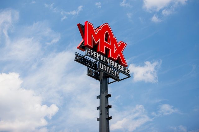 Sign displays "MAX PREMIUM BURGERS DRIVE IN" with red and white letters, standing tall against a blue sky with clouds.