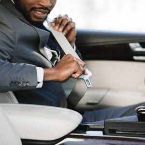 Man fastening seatbelt inside a car, dressed in a suit, with a steering wheel and interior details visible.