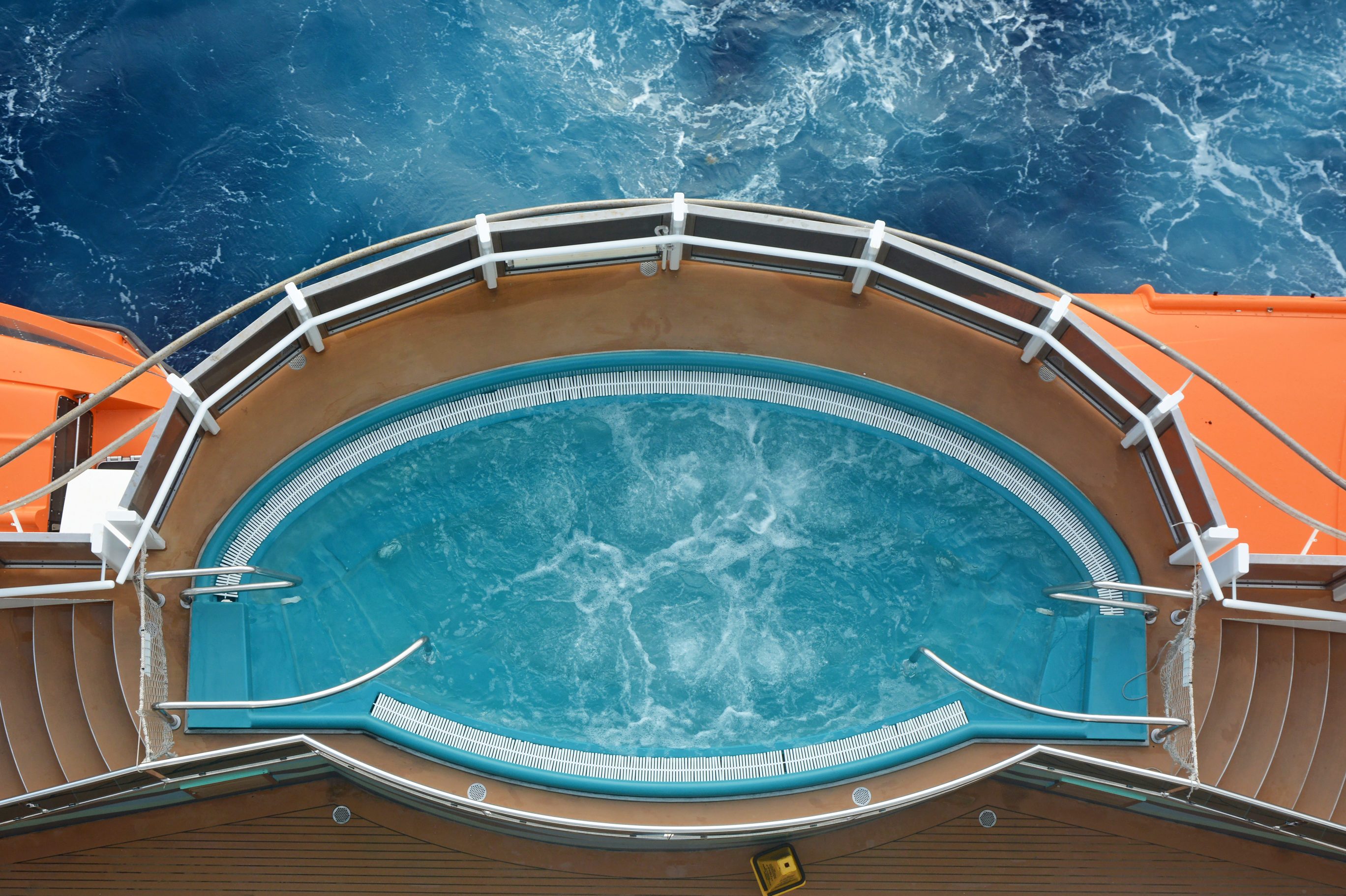 Oval pool with swirling water, enclosed by white railings. Orange structure and ocean waves surround the deck area.