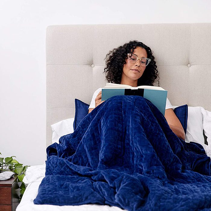 Person reads book under a blue blanket in bed, surrounded by pillows with a beige headboard. A nightstand with a plant is nearby.