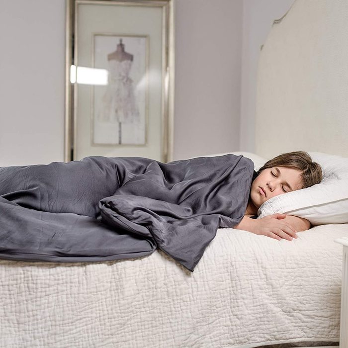 Person sleeping under a dark gray blanket on a quilted bed; a framed dress photo hangs on the wall in the background.