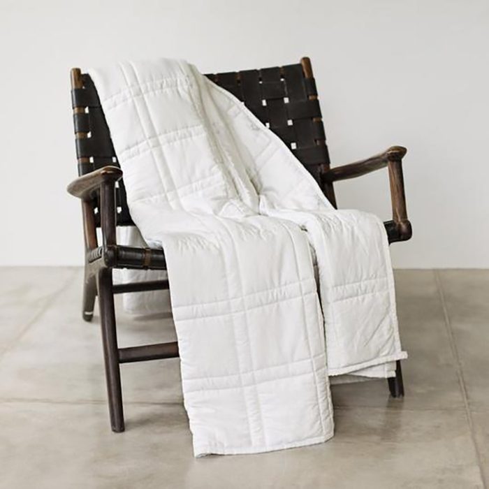 A white quilt drapes over a dark wooden armchair, positioned on a light-colored tiled floor against a plain wall.