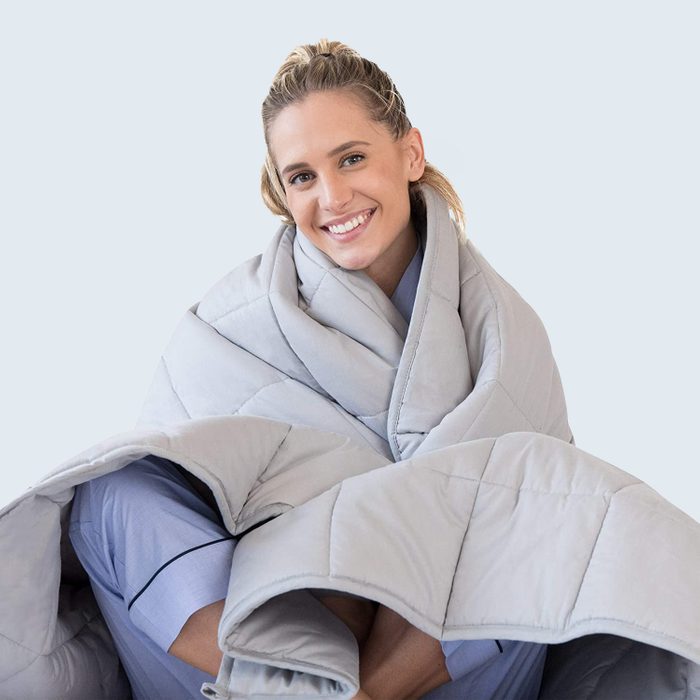 Person smiling, wrapped in a gray quilted blanket, sitting in a light, neutral background.