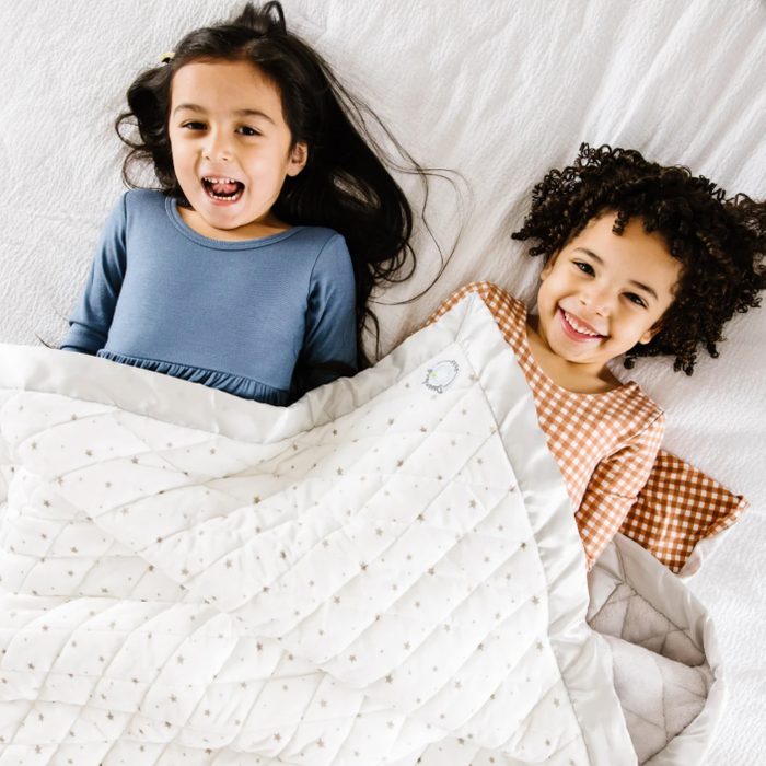 Two children laughing, lying under a white quilt with star patterns on a bed with white sheets.