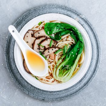 A bowl contains noodles with shiitake mushrooms, bok choy, and sesame seeds; a spoon rests on top, within a minimalist stone setting.