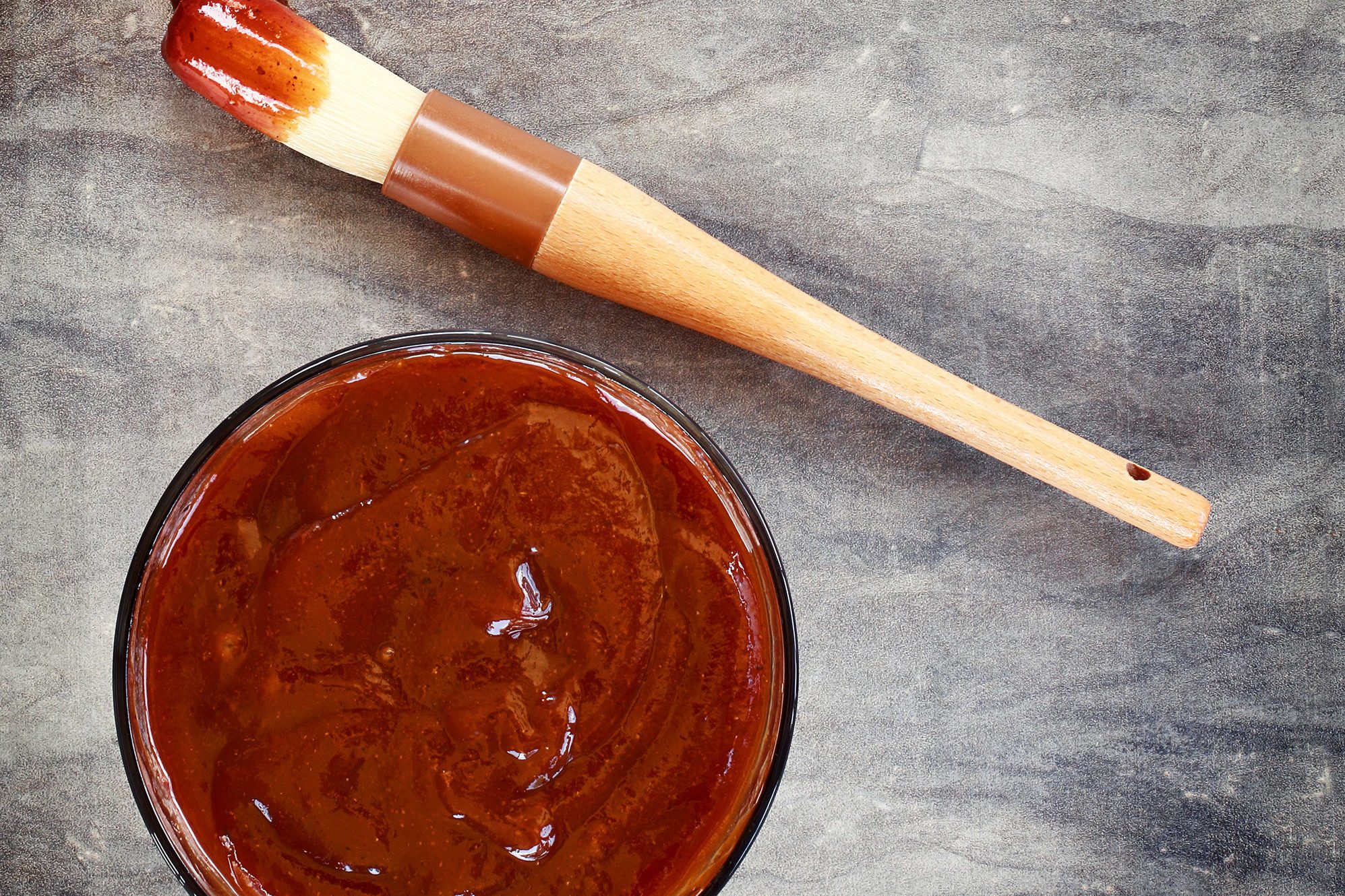 Barbecue sauce fills a glass bowl, sitting on a textured surface, with a sauce-covered basting brush lying beside it.