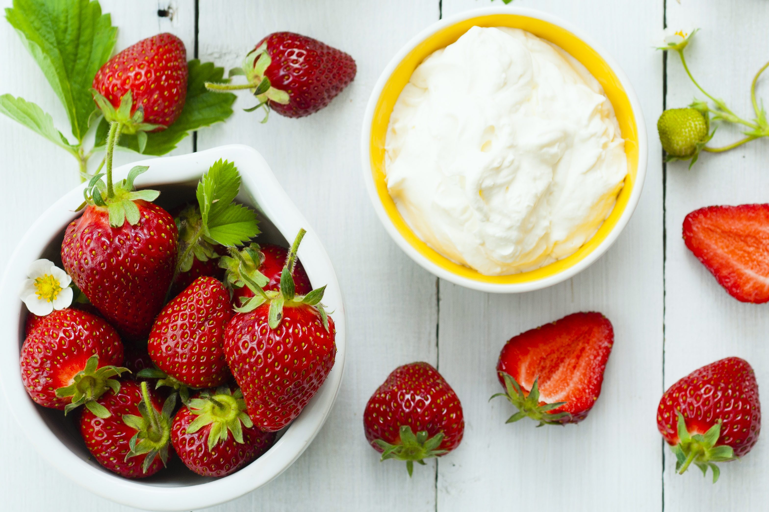 Strawberries rest in a bowl, surrounded by sliced pieces and a dish of cream, on a white wooden surface.