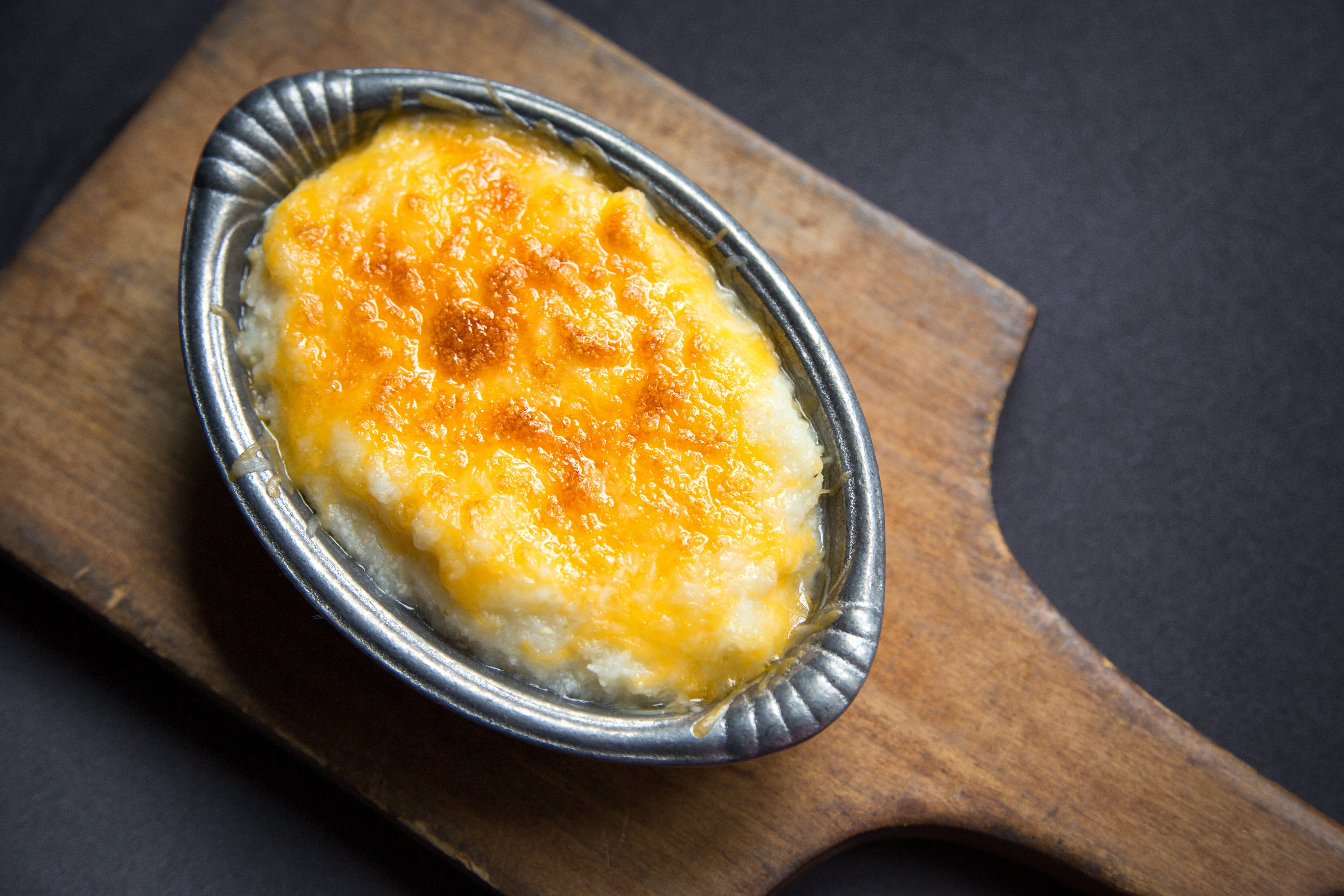 A baked cheese dish with golden crust sits in a small metal dish on a wooden board, on a dark background.