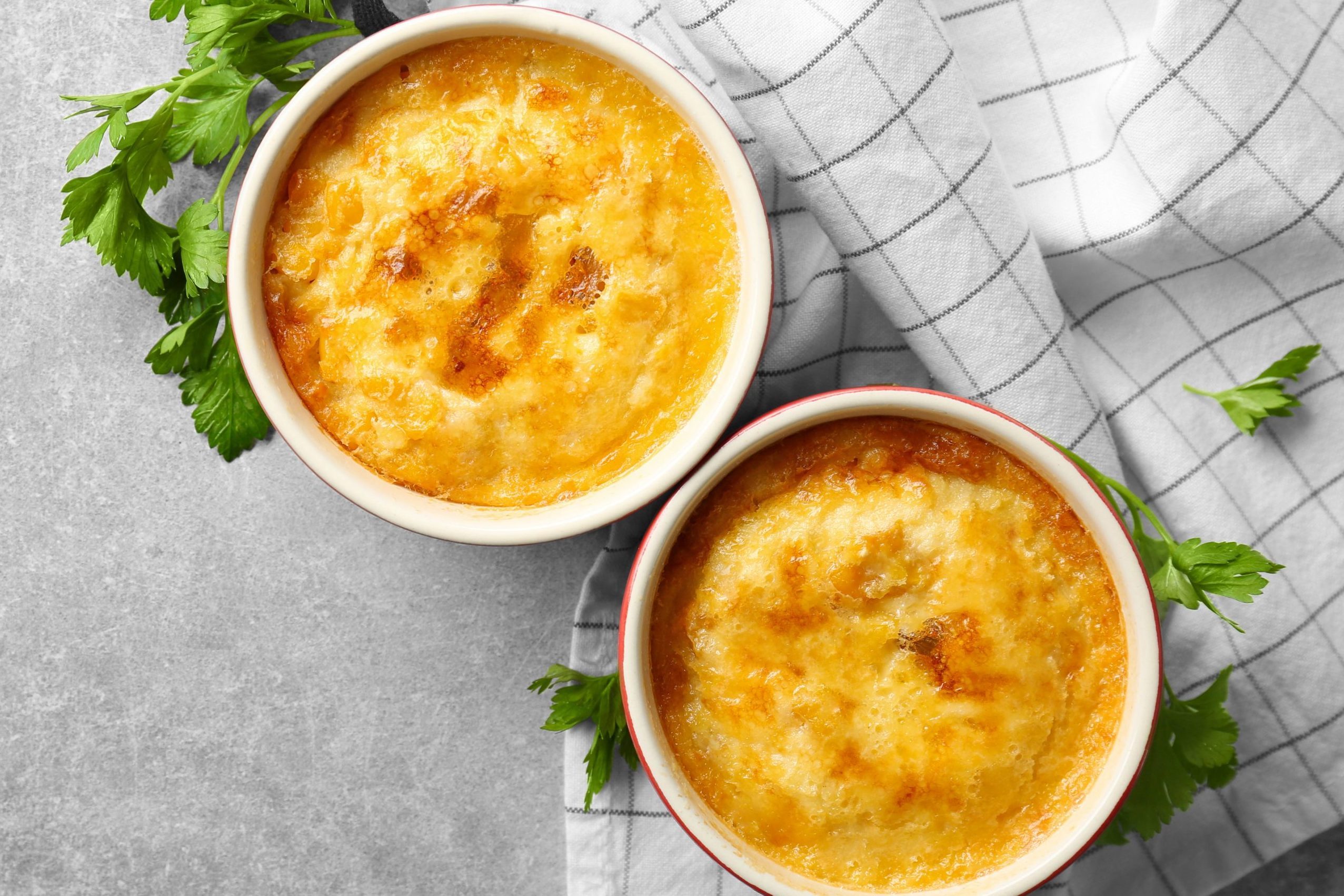 Two baked dishes, bubbling and golden, sit on a checkered cloth with parsley garnish on a gray tabletop.