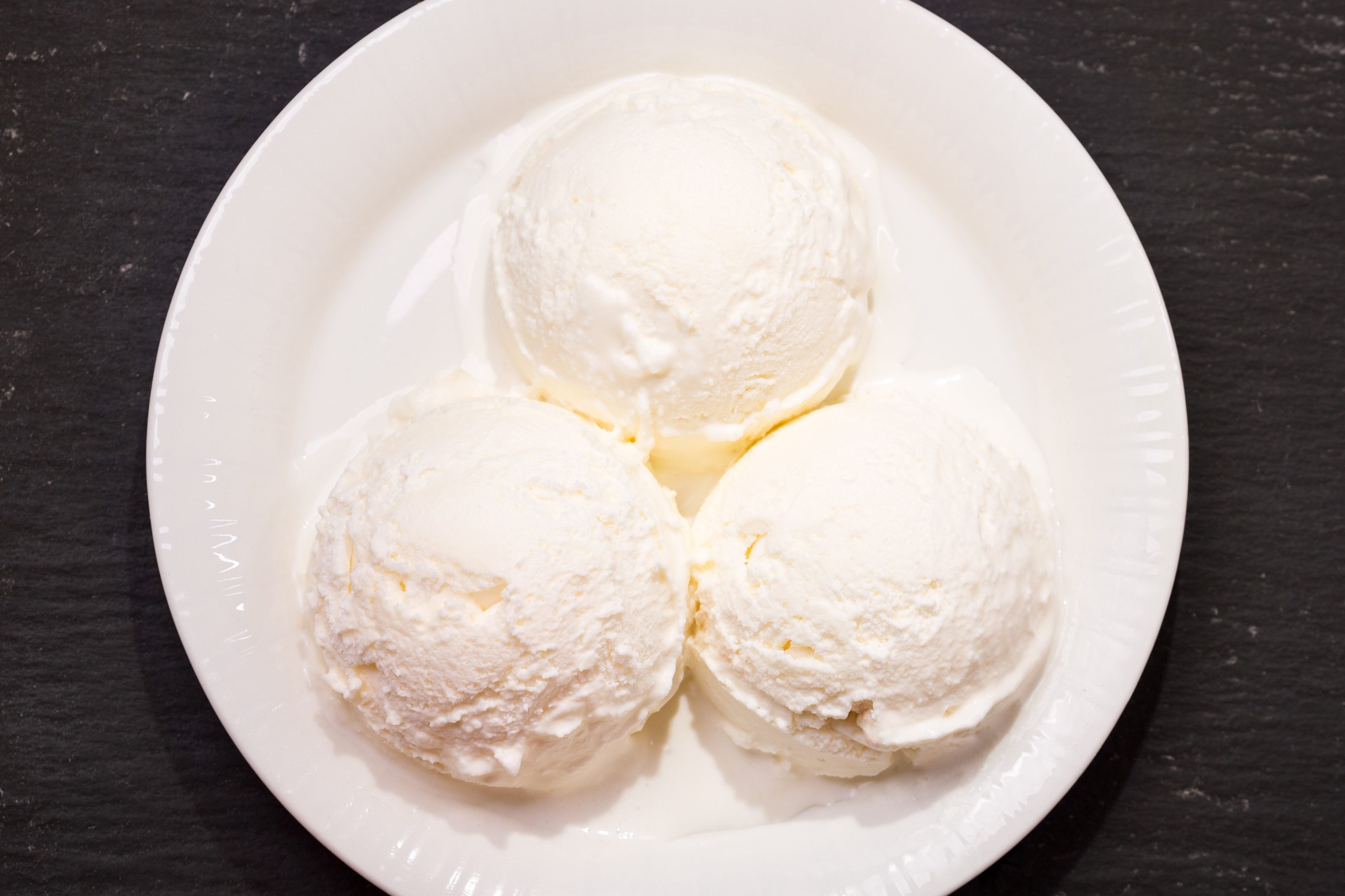 Three scoops of vanilla ice cream sit on a white plate against a dark, textured background.