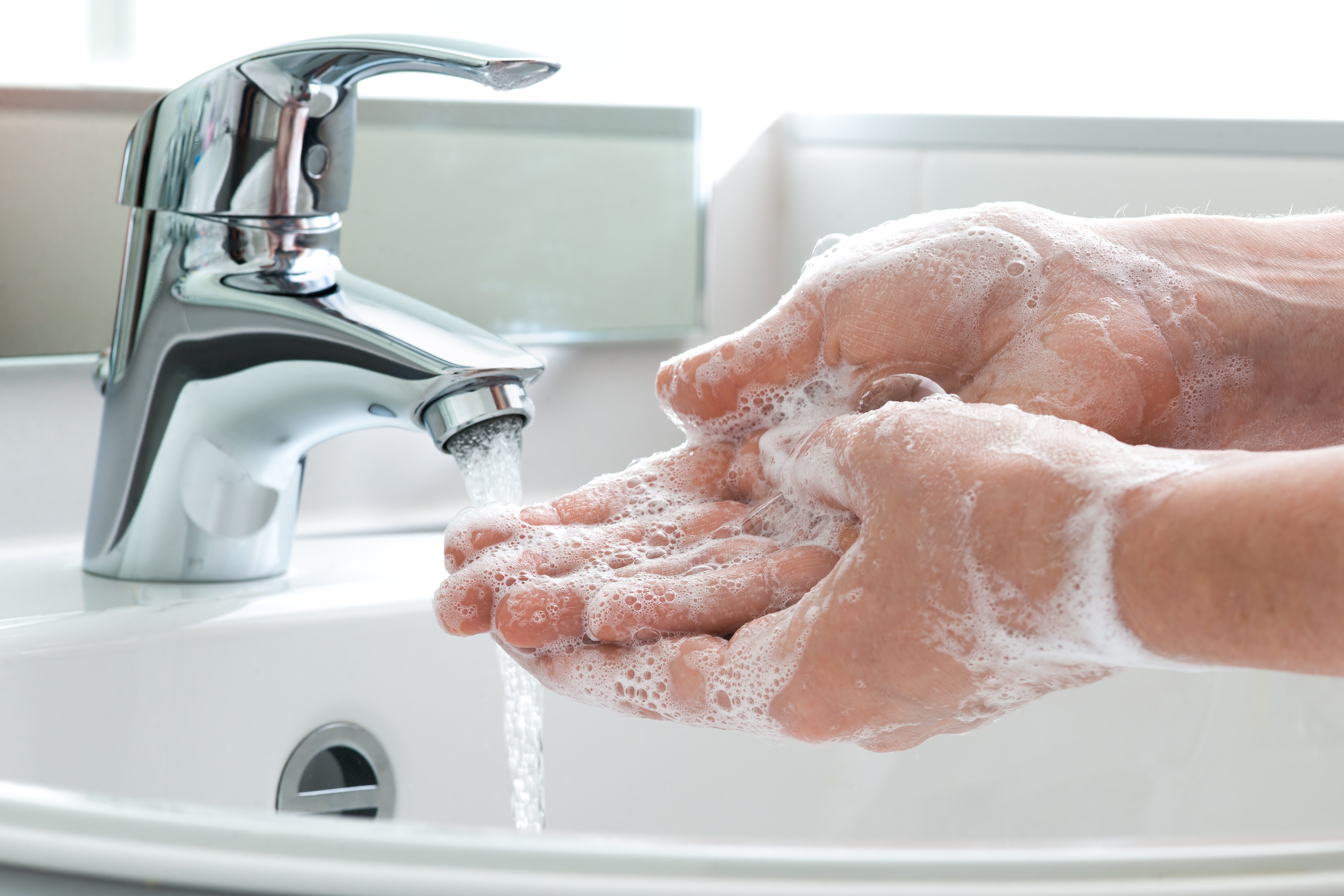Hands covered in soap lather are being washed under a running faucet in a bathroom sink.