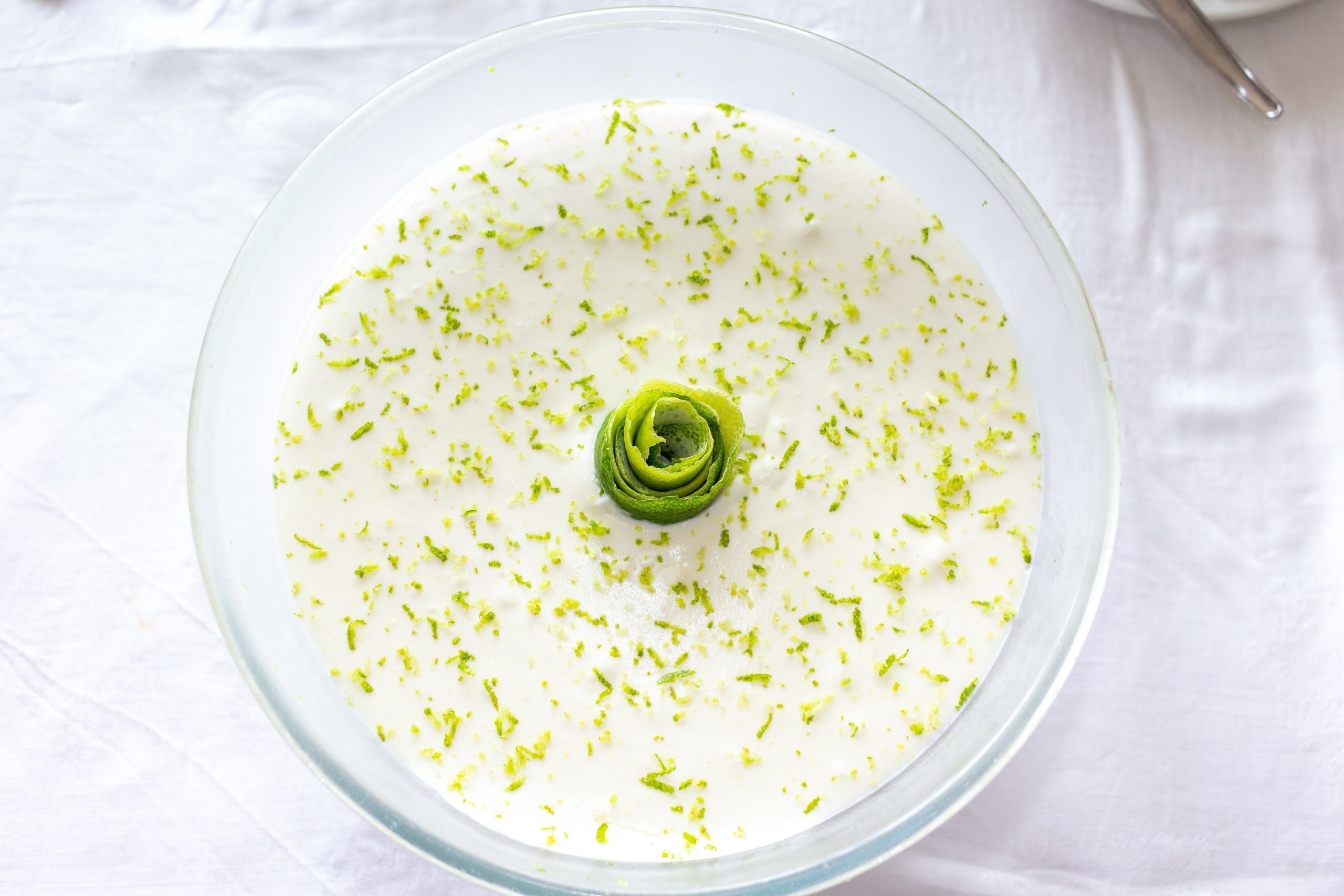 Dessert in a glass bowl, topped with lime zest and a lime spiral, on a white tablecloth.
